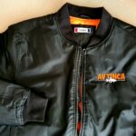 Schwarze Bomberjacke mit orangefarbenem Innenfutter und dem Schriftzug AVTINCA mit weißer Grafik auf der linken Brust, flach auf einer Fläche ausgestellt.