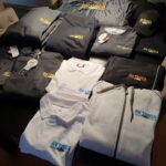 Verschiedene Markenartikel der AVTNCA, darunter Polohemden, Kapuzenpullis, T-Shirts, Mützen und Jacken, liegen ordentlich auf einer ebenen Fläche.